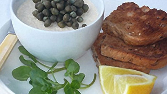 a picture of a small white bowl containing potted haddie with capers on top. sitting on a larger plate with slices of bread a wedge of lemon and some watercress on the side
