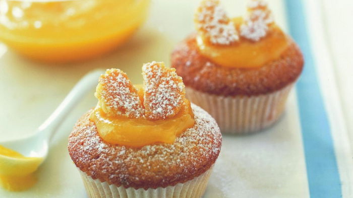 Two cupcakes topped with powdered sugar and slices resembling butterfly wings, placed on a white surface. A bowl of orange sauce and a spoon are in the background.