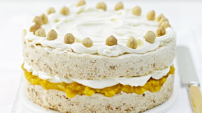 A layered cake with icing topped with whole hazelnuts. The middle layer contains a yellow filling, possibly fruit-based, creating a contrast with the beige cake. The cake is presented on a white surface with a knife beside it.