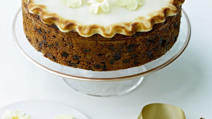 An Easter Simnel Cake on a glass cake stand decorated on the top with icing and little yellow and white flowers, next to a white plate of more white and yellow flowers and a gold ribbon next to it