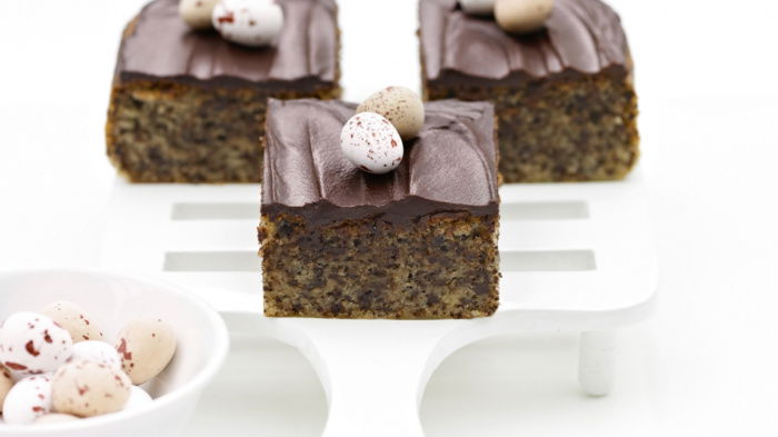 Three chocolate-topped cake squares on a white serving spatula, each decorated with small speckled mini eggs. A bowl of additional eggs is in the foreground on a white surface.