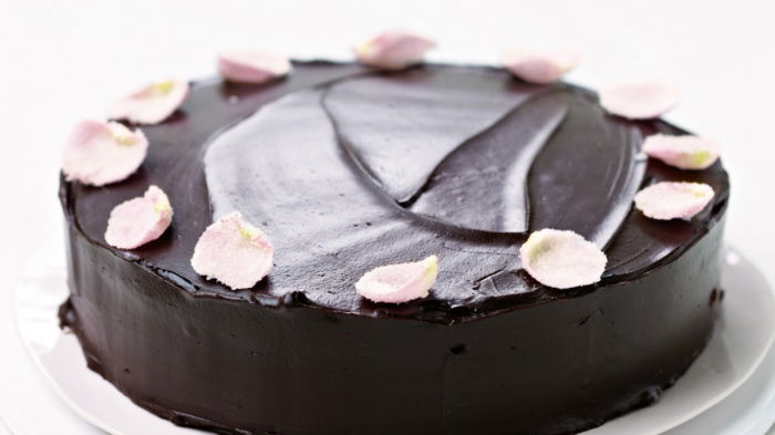 A round chocolate cake with smooth, dark frosting on a white platter. The cake is decorated with light pink flower petals around the edge, with a few petals scattered on the platter. The background is white.