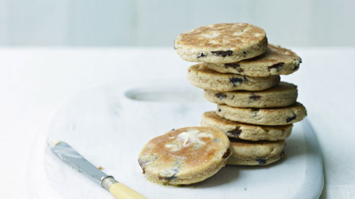 A stack of toasted cakes with raisins sits on a white marble board. A small butter knife with a light yellow handle rests beside the stack. The background is a soft white, creating a minimalist and airy setting.