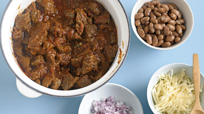 A bowl of beef chilli in a white saucepan with little bowls of grated cheese, chopped onion, beans and cream surrounding it