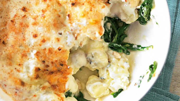 An aeiral view of a white bowl of cooked gnocchi, spinach and dolcelate sauce mixed up together with a portion removed, sitting on a folded blue napkin