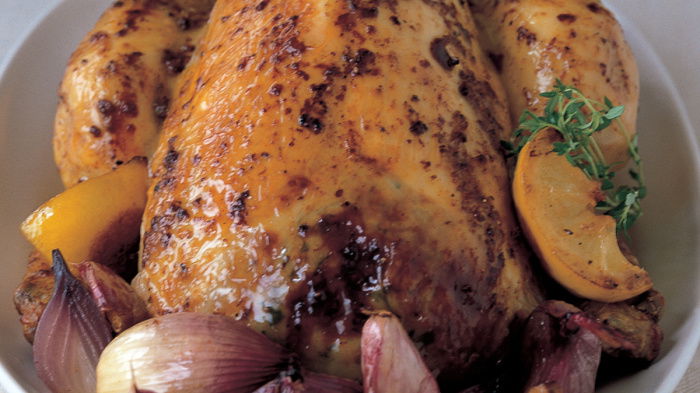 A picture of a roasted chicken in a dish surrounded by lemon wedges, thyme and garlic