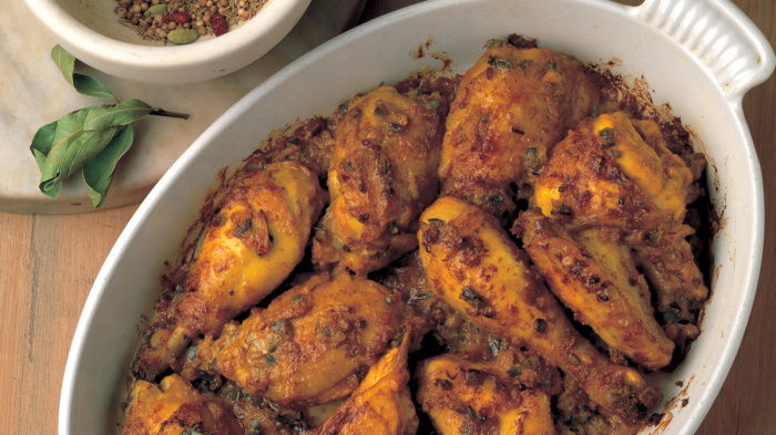 A white oval dish with pieces of spiced cooked chicken, a pestle and mortar next to it with ground spices and a plate of rice
