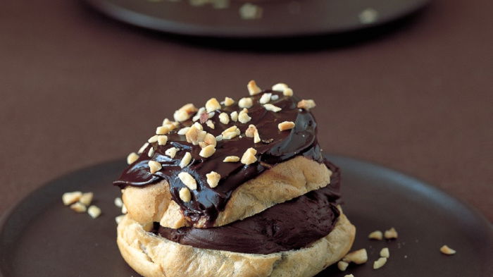 A choux bun split in half horizontally and filled and topped with a chocolate mousse mixture and coated with chocolate icing and chopped nuts.