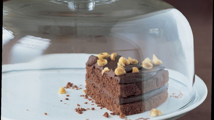 A slice of chocolate cake on a white cake stand with a glass lid