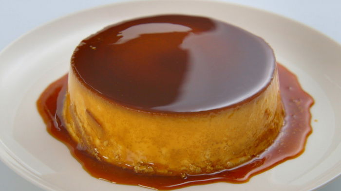 A smooth, glossy caramel flan sits on a white plate, its rich caramel sauce dripping down the sides and pooling around the base. The dessert has a golden-brown top and a creamy, custard-like texture.