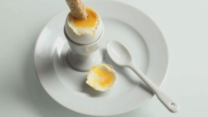 a white china egg cup on a white plate, with a soft boiled egg opened and a solder being dipped in the yolk