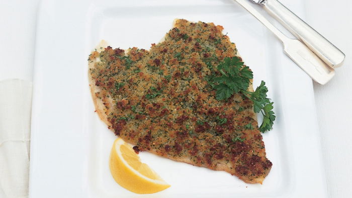 A picture of a fillet of roasted fish and a wedge of lemon on a white plate with a knife and fork resting on the side and two little silver pots of salt and pepper