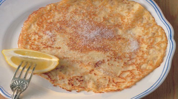 A picture of a cooked pancake covered in sugar on a white plate with a wedge of lemon on a fork resting on the side