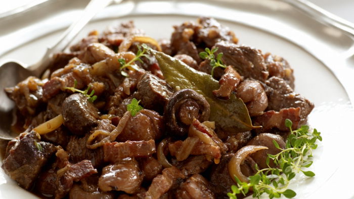 A white plate of cooked slices of venison, mushrooms and bacon in a sauce with some garnish on the side and a spoon resting on the rim