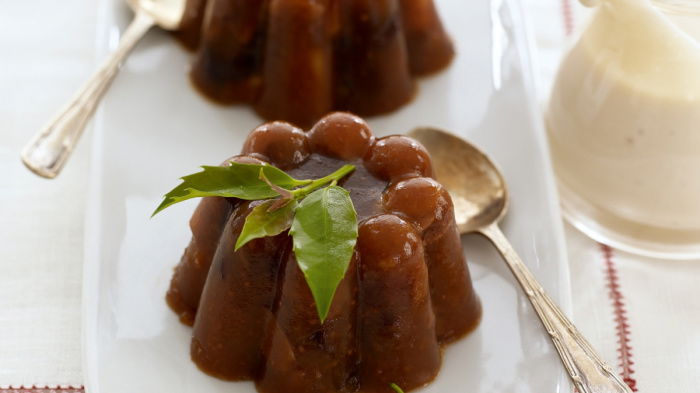 two pretty chrismas puddings on a whilte place with a spoon resting on the side and a jug of cream