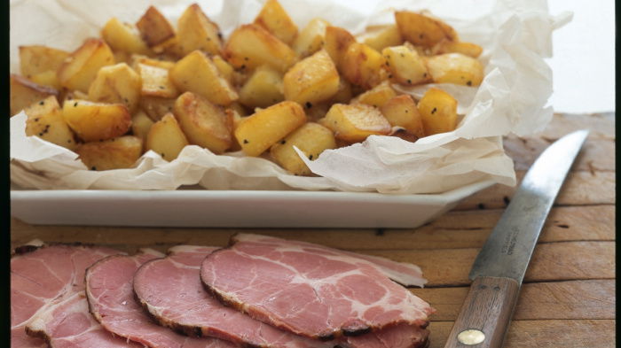A picture of diced sauteed potatoes in a white dish sitting on a wooden board with sliced ham and a knife on the side
