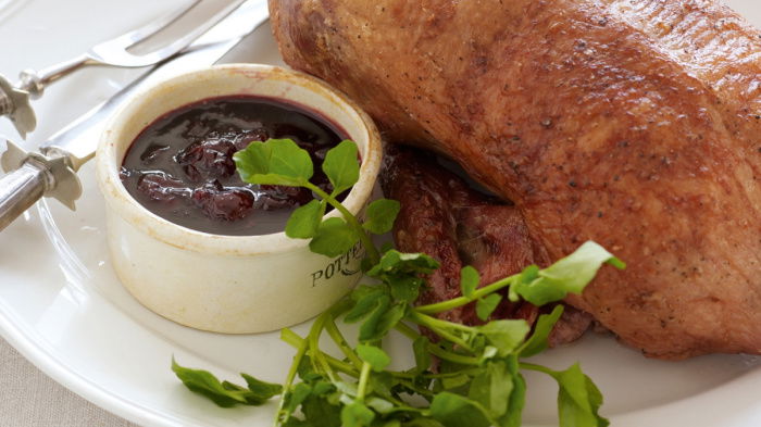 a picture of a white plate with a whole roast duck with a bowl of cherry sauce and a silver carving knife and fork