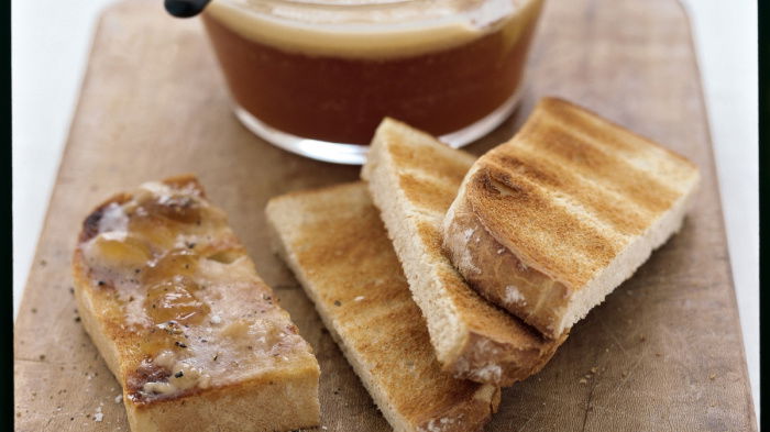 A picture of sliced toast on a wooden board with a glass container of turkey dripping and a knife resting on the edge