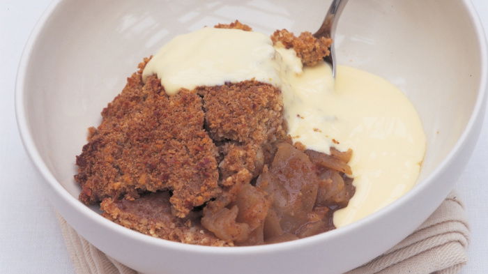 A white bowl of apple crumble with custard poured over the top and a spoon resting on the side