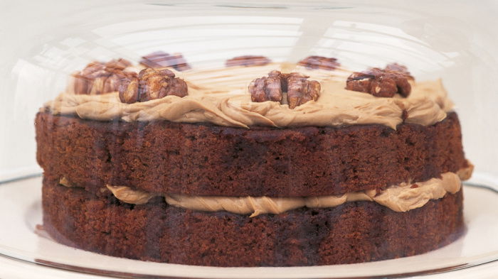 A layered cake with frosting and walnuts on top, displayed under a glass dome.