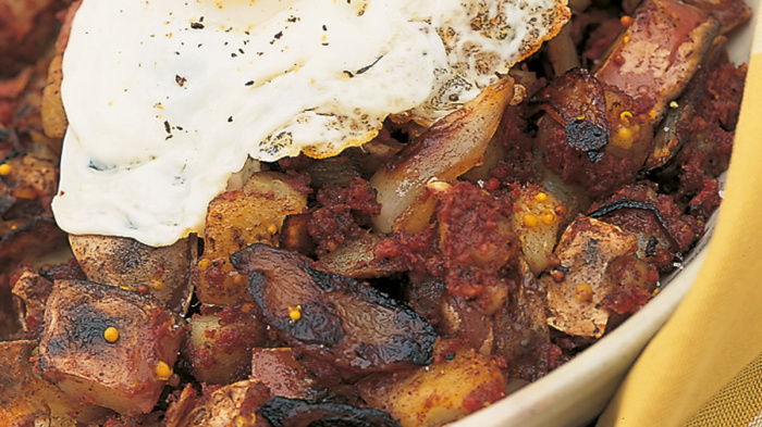 A plate of crispy corned beef hash topped with a sunny-side-up egg. The hash includes diced potatoes and onions, with a golden-brown crust. A yellow and white checkered cloth is partially visible beneath the plate.