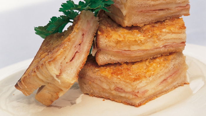 Stacked croque monsieur sandwiches on a white plate, garnished with parsley. The sandwiches have layers of melted cheese and ham, with a golden, crispy crust.