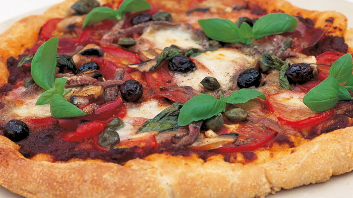 A cooked pizza on a white plate with a topping of basil, tomato, cheese and olives