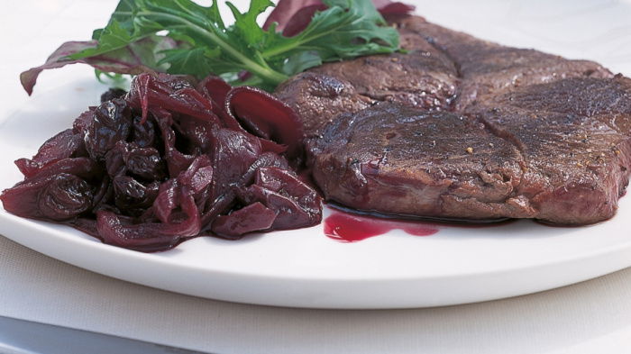 A grilled venison steak on a white plate with confit over the top and a garnish
