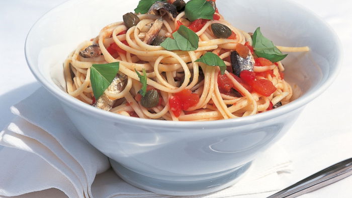 A white bowl of linguine pasta with tomatoes, capers, sardines and some torn basil to garnish. A fork is next to the bowl