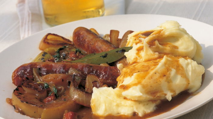 A picture of braised sausages with a pile of fluffy mash on a white plate with a glass of cider in the background