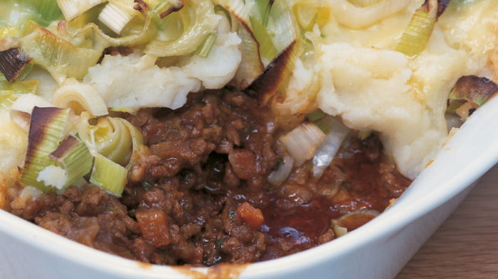 A white dish of shepherds pie with a portion removed showing minced lamb and vegetables and a topping of mashed potato, leeks and cheese