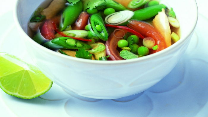 A white soup bowl on a matching plate containing hte borth with different vegetables floating on the top and a wedge of lime on the plate