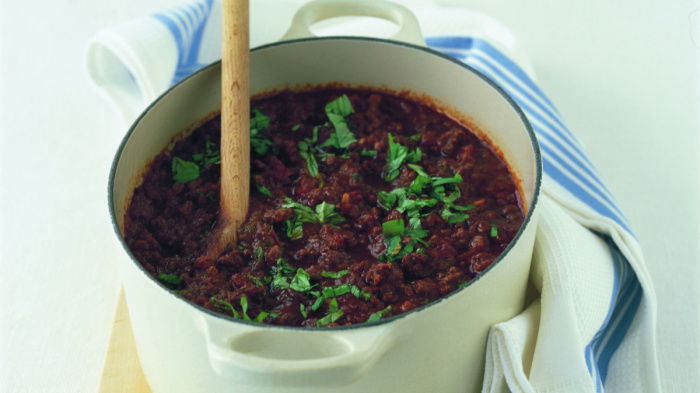 a white casserole dish containing ragu bolognaise sauce with basil sprinkled on top, and a blue and white tea towel