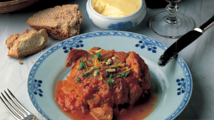 A picture of a blue and white china plate containing coked ossobuco with a knife and fork a glass of red wine and some bread and butter on the side