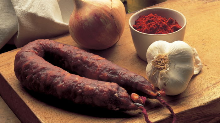 Onion, garlic, chorizo sausage and a cup full of paprika sat on a wooden chopping board