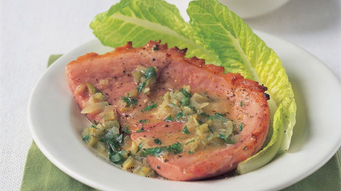 A slice of grilled gammon topped with a gherkin and white wine sauce, with romaine lettuce on a white plate, placed on a green napkin. A small bowl of sauce is in the background.