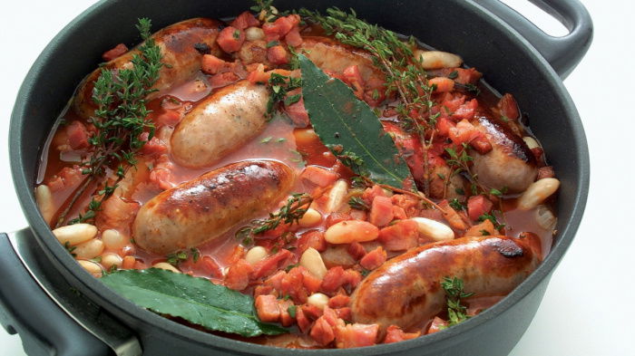 A picture of cooked cassoulet in a black casserole showing sausages, beans, bay leaves and tomatoes