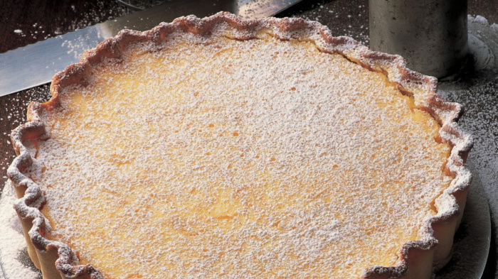 A round lemon tart on a dark plate, dusted with powdered sugar. The tart has a fluted edge. In the background, there is a knife, a sifter, and a small bowl of cream. The dark surface is sprinkled with sugar.