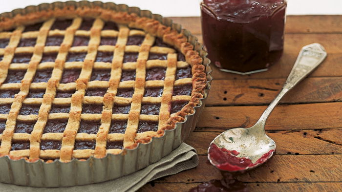 a picture of a linzertorte tarte in a flan tin with latticed pastry across the top and a jar of jam and spoon next to it