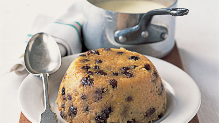 A picture of the pudding turned out on to a white dish with a serving spoon resting on the side and a pan of sauce in the background