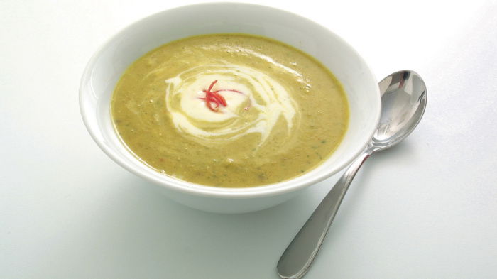 A bolw of chickpea, chilli and coriander soup in a white bowl with a swirl of creme fraiche and garnish of chilli on top and a soup spoon on the side