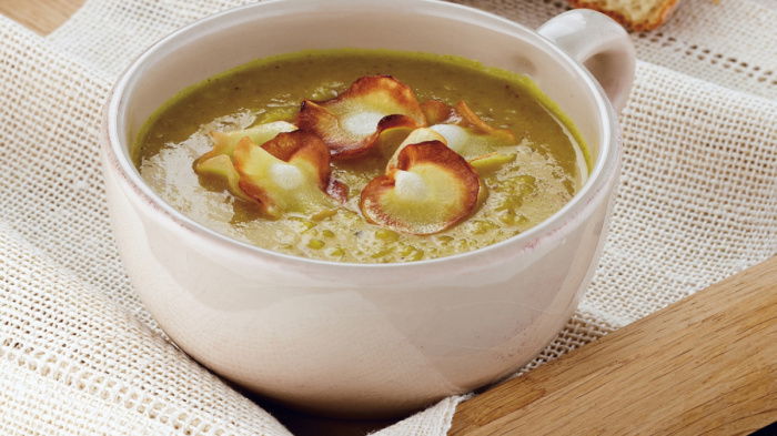 A bowl of parsnip soup in a white mug, with parsnip crisps on the top, sittin on a wooden tray with a roll of white bread next to it