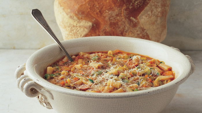 A white bowl with pasta soup garnished with herbs and cheese, with a spoon inside. A round loaf of bread is placed behind the bowl on a marble surface.