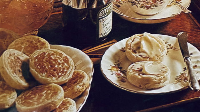 a table set with a china cup of tea and a saucer, a side plate with two muffins covered in butter, a plate of muffins, a jar of jam, a dish of marmalade and a roaring fire in the background