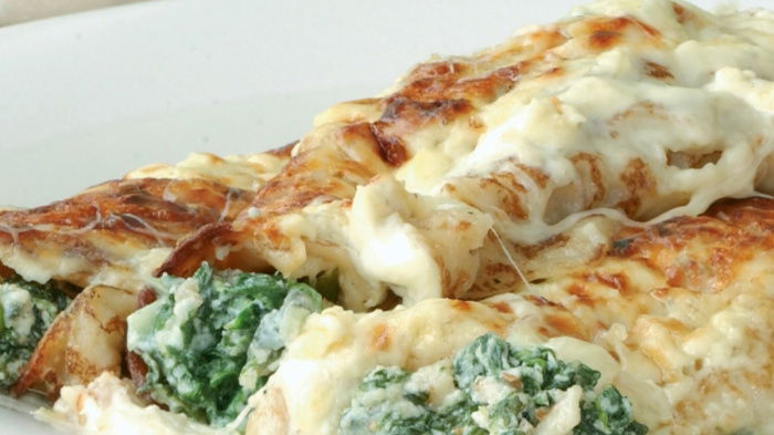 A picture of cooked pancake cannelloni on a white plate with spinach and a cheese sauce