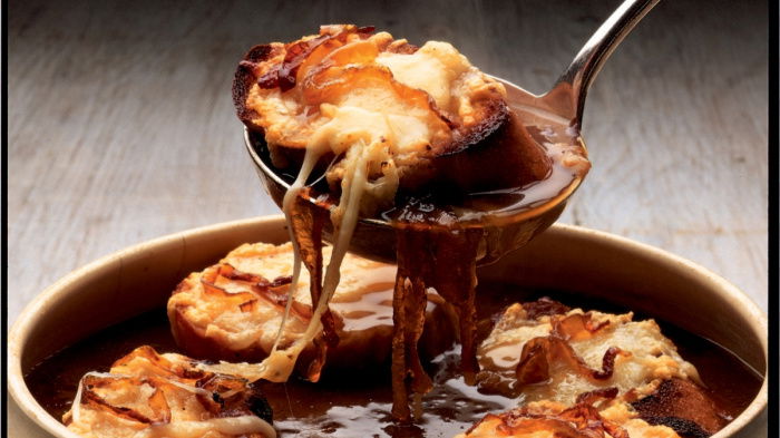 A picture of a soup bowl full of french onion soup with croutons covered in melted cheese and a ladle lifting out a whole crouton