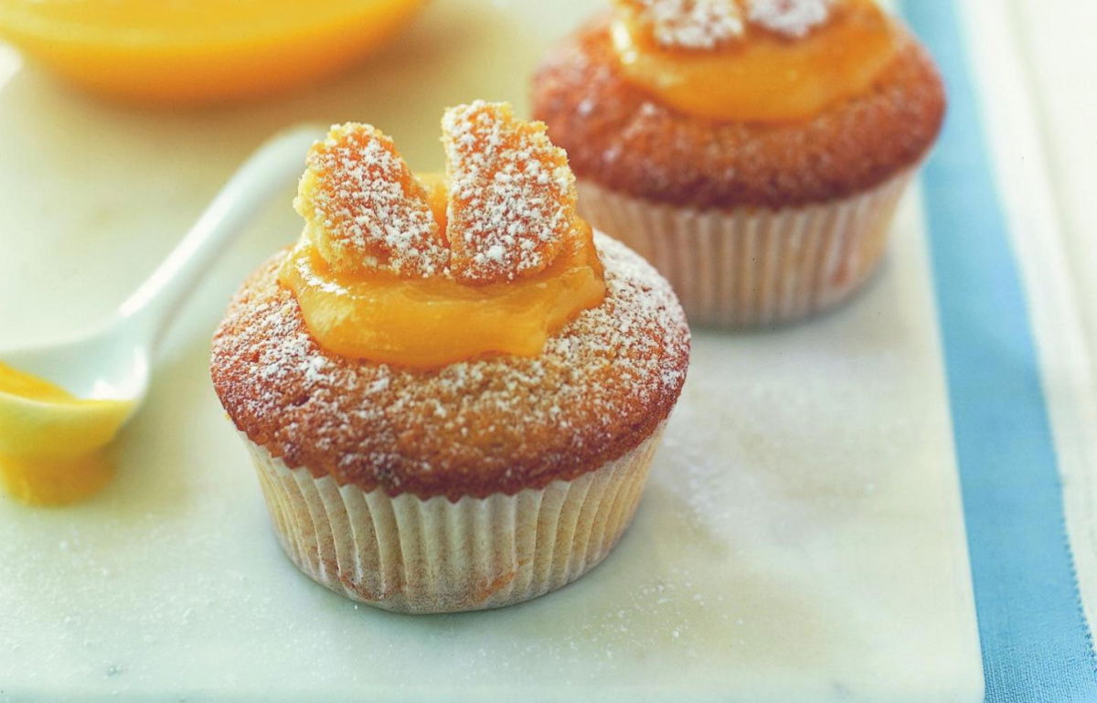 Two cupcakes topped with powdered sugar and slices resembling butterfly wings, placed on a white surface. A bowl of orange sauce and a spoon are in the background.