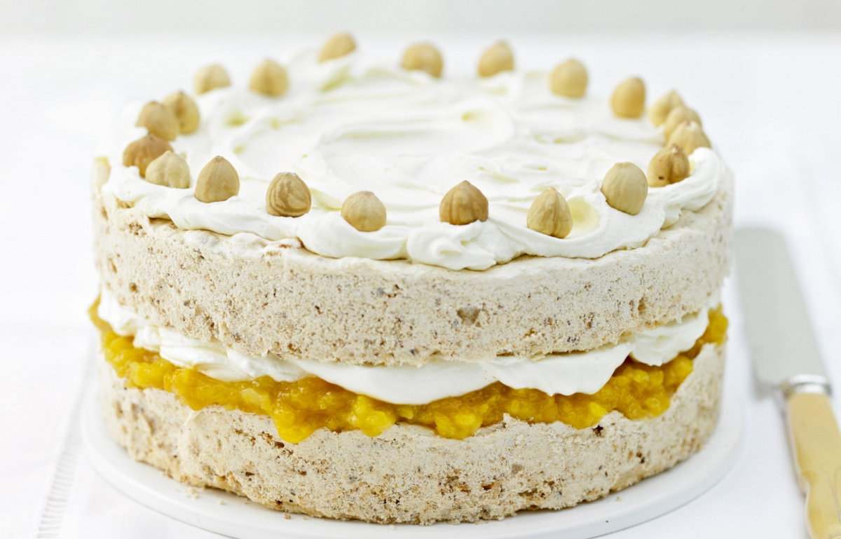 A layered cake with icing topped with whole hazelnuts. The middle layer contains a yellow filling, possibly fruit-based, creating a contrast with the beige cake. The cake is presented on a white surface with a knife beside it.