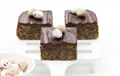 Three chocolate-topped cake squares on a white serving spatula, each decorated with small speckled mini eggs. A bowl of additional eggs is in the foreground on a white surface.