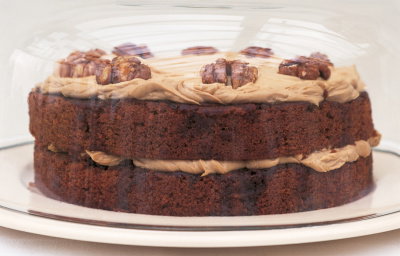 A layered cake with frosting and walnuts on top, displayed under a glass dome.